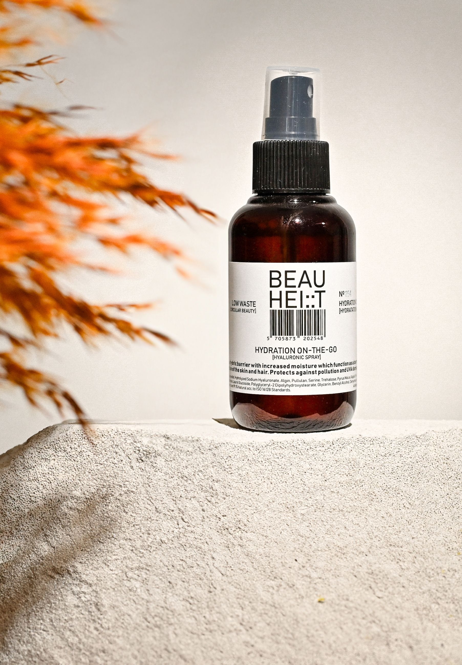 Braune Sprühflasche mit schwarzem Sprühkopf. Aufschrift: BEAUHEIT, HYDRATION ON-THE-GO, Hyaluronsäure Spray. Auf Stein platziert, mit Pflanzen im Hintergrund.