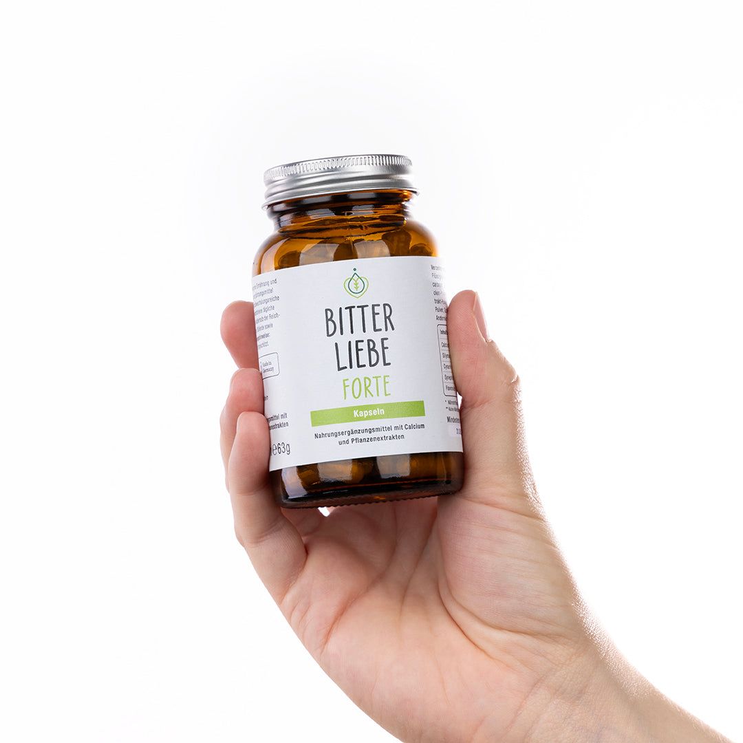 Hand hält eine braune Glasflasche mit "BitterLiebe Forte". Silberner Deckel, weißes Etikett.