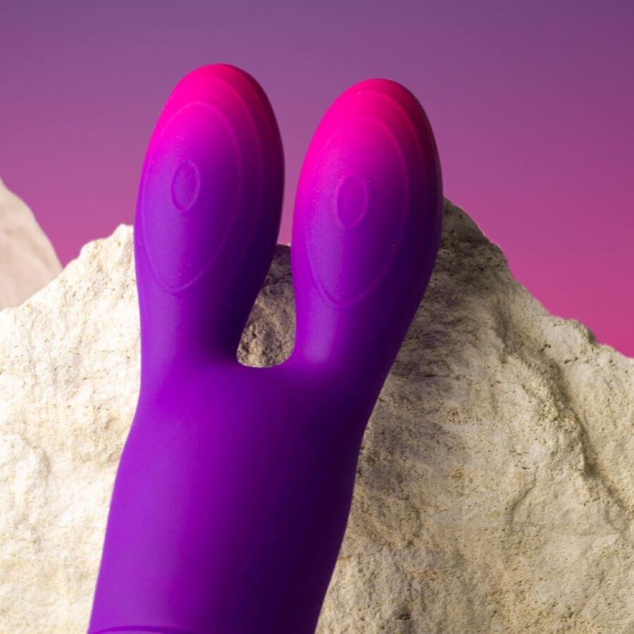 Lila Vibrator mit Hasenohren-Design. Farbverlauf von Lila zu Pink. Auf Stein platziert, vor rosa-violettem Hintergrund.