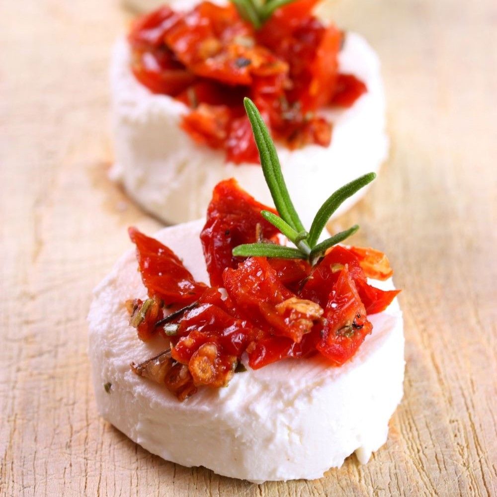 Zwei runde Käse mit getrockneten Tomaten belegt. Rosmarinzweig als Garnitur. Auf Holzuntergrund.