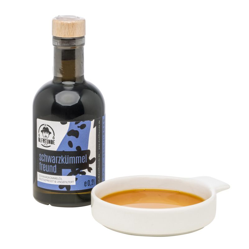 Braune Glasflasche mit Holzverschluss und Schale mit Flüssigkeit. Etikett mit Produktname und Logo. Schwarzkümmelöl.