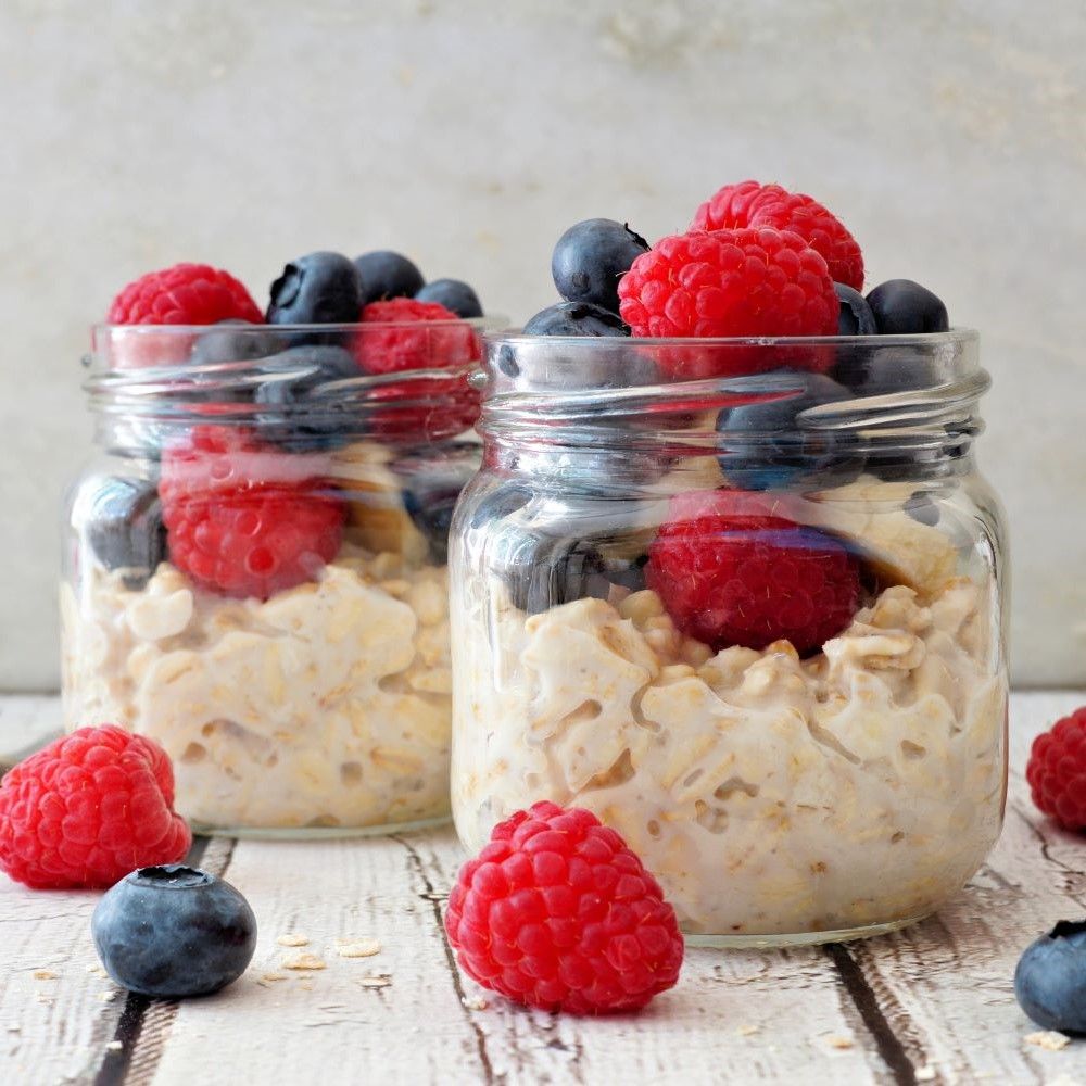 Zwei Gläser Porridge mit Himbeeren und Blaubeeren. Auf Holzoberfläche platziert.