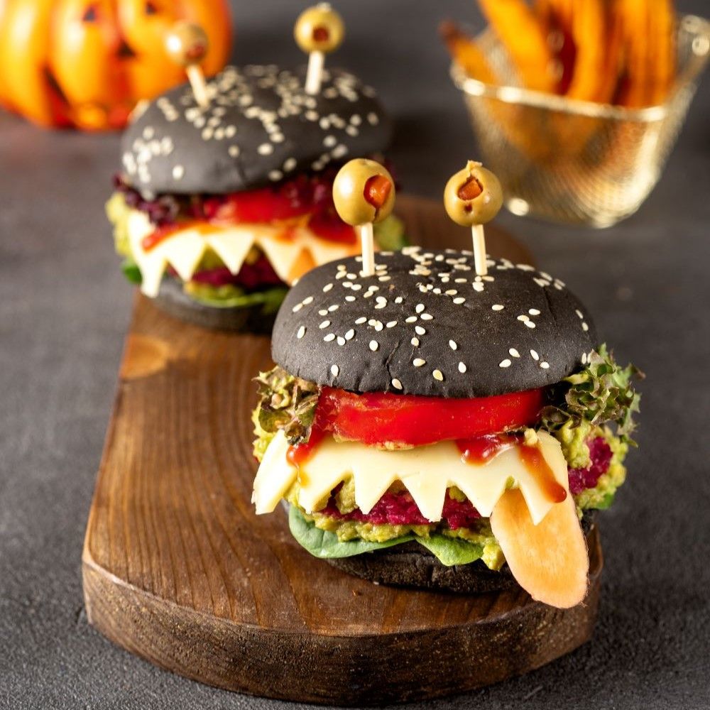 Zwei Halloween-Burger auf Holzbrett. Schwarze Brötchen, Oliven-Augen, Käse-Zähne. Burger mit Gemüse gefüllt.