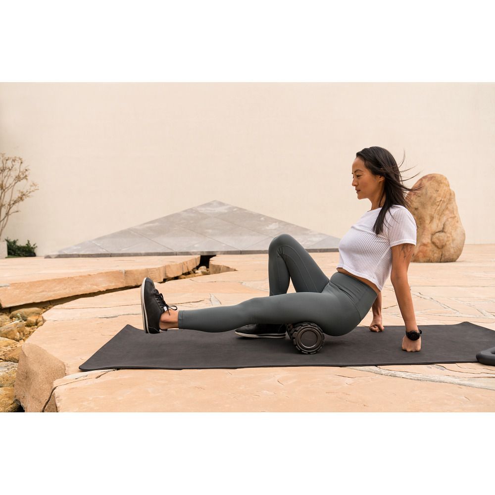 Frau sitzt auf einer Matte und verwendet eine Vibrationsmassage-Rolle am Bein.