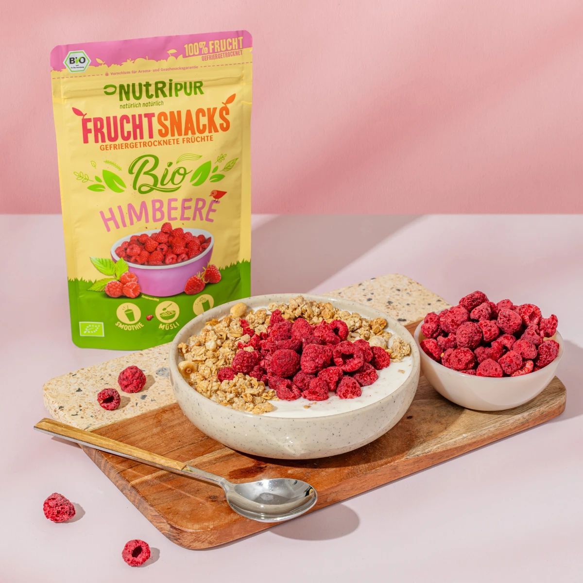 Beutel und Schalen mit gefriergetrockneten Himbeeren. Schale mit Müsli und Himbeeren. Holzbrett und Löffel.