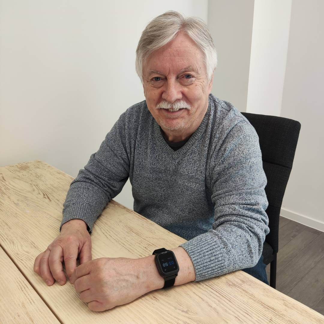 Älterer Mann mit grauer Strickjacke sitzt am Tisch, trägt eine schwarze Smartwatch.
