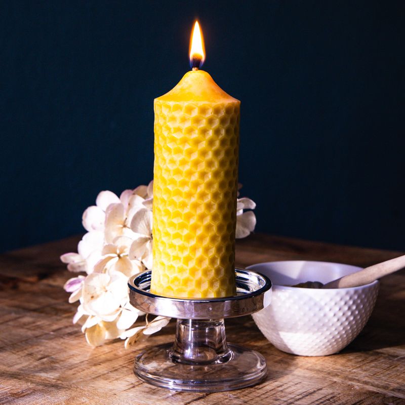 Gelbe Wachskerze mit Wabenmuster, brennend. Auf einem silbernen Kerzenhalter, neben einer Schale und Blumen auf Holz.