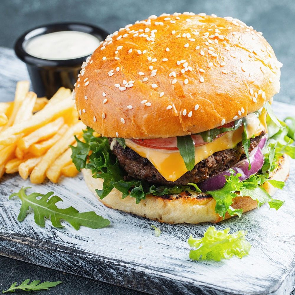 Burger auf Holzbrett. Brötchen mit Sesam, Fleisch, Käse, Salat, Tomate, Zwiebel. Pommes und Sauce daneben.