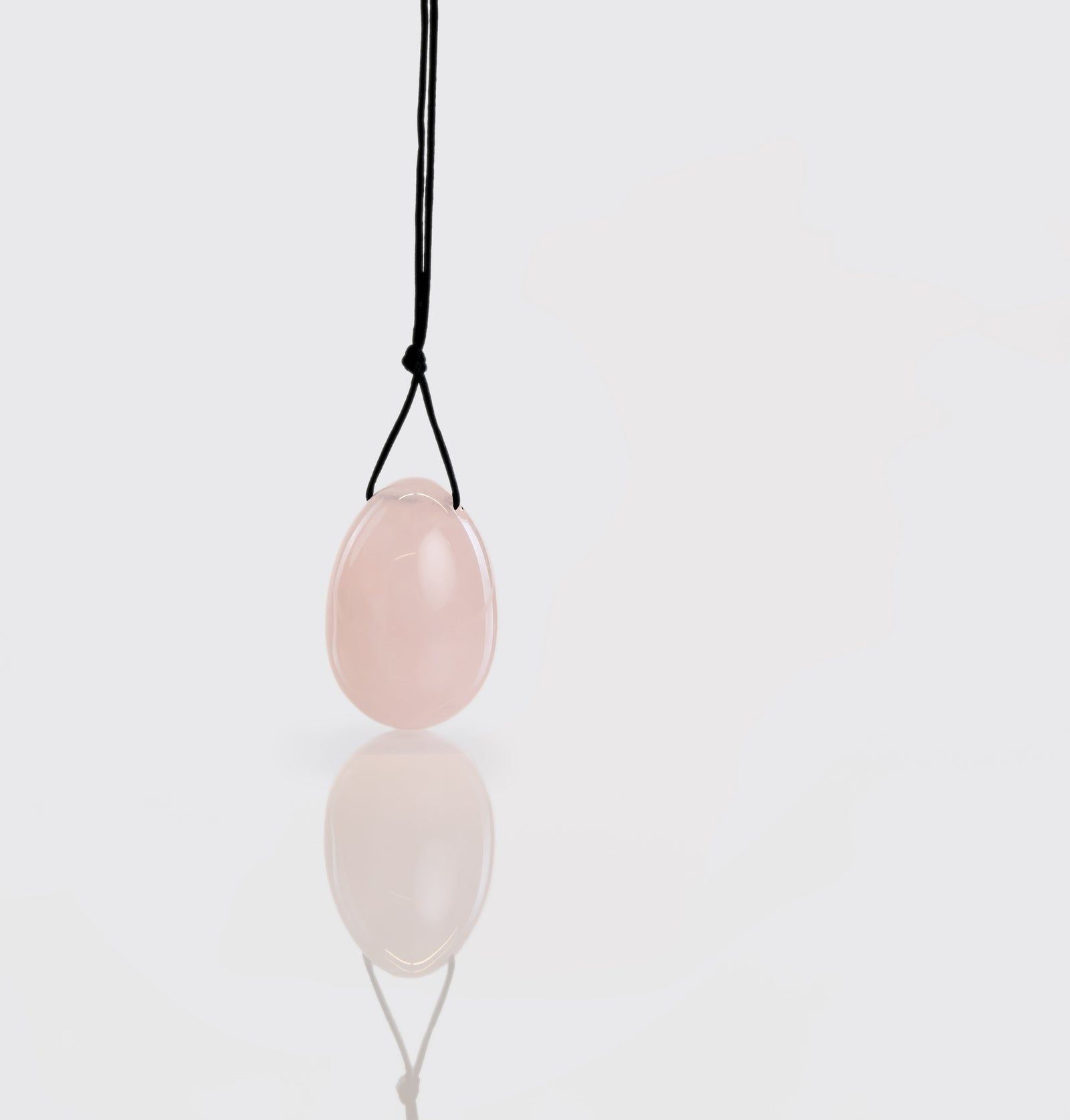Rosafarbenes Yoni-Ei mit schwarzer Schnur, auf glänzender Oberfläche. Spiegelung des Eis sichtbar.
