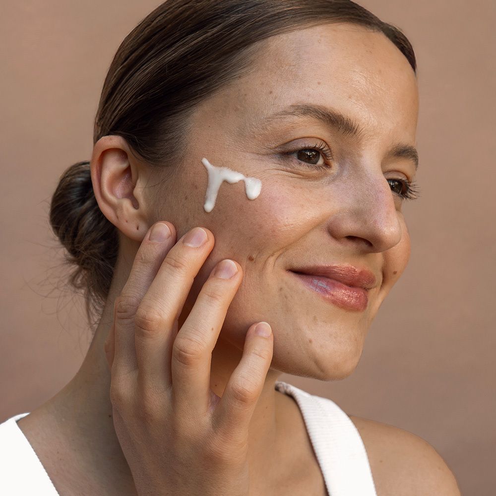 Frau mit Serum auf der Wange. Lächelt. Text: Natürliche, ohne Duftstoffe. Natürlicher Eigengeruch des Bakuchiols. Ohne Duftstoffe für mehr Hautverträglichkeit.