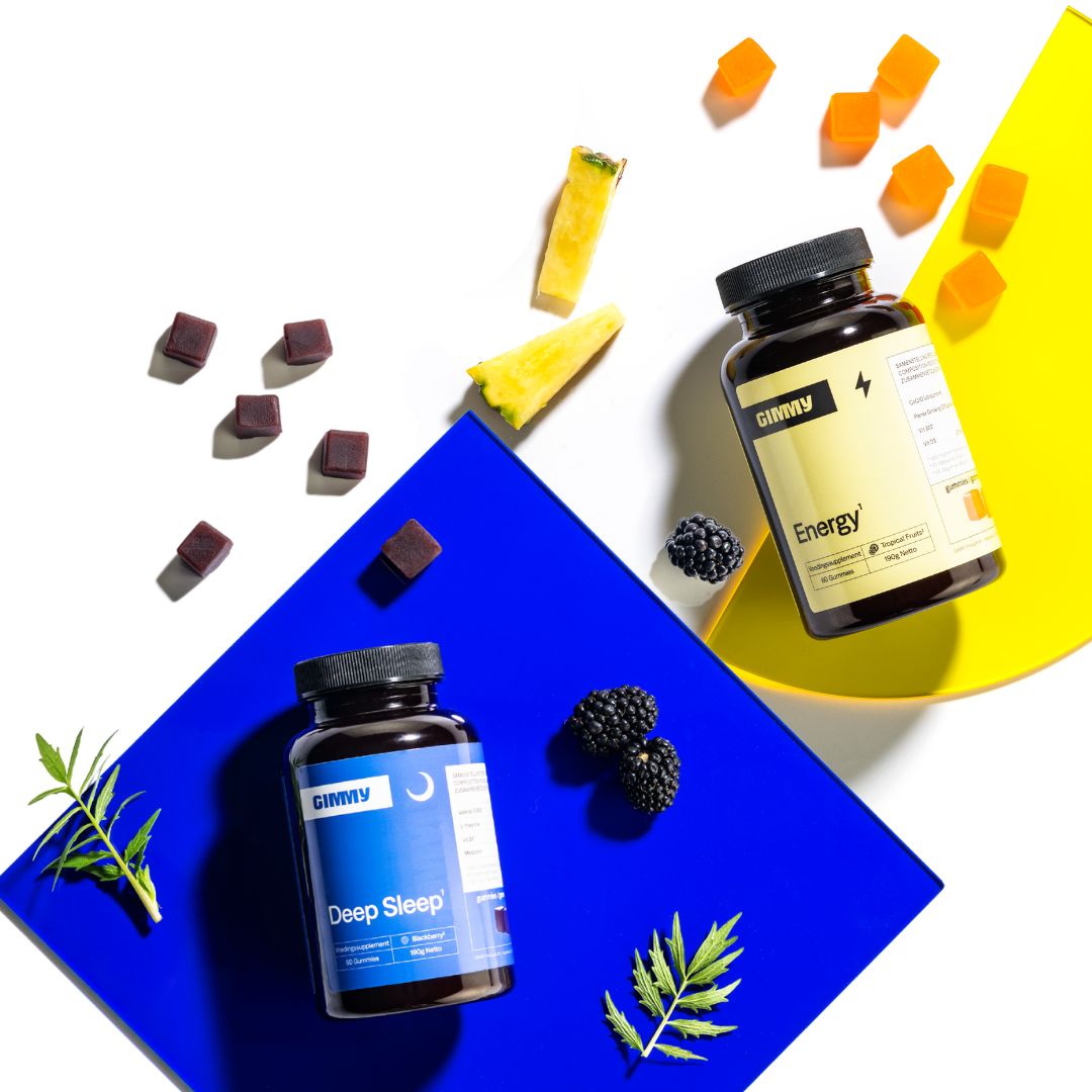 Zwei Flaschen mit schwarzen Deckeln. Eine gelbe Etikett mit "Energy", eine blaue mit "Deep Sleep". Gummibärchen und Früchte daneben.