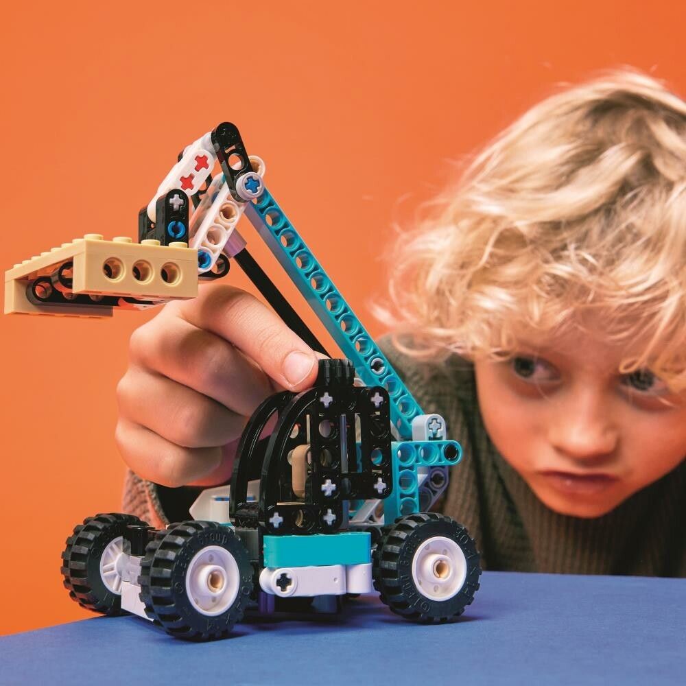 Un bambino guarda un modello di sollevatore telescopico LEGO Technic completato. Il modello ha elementi blu, neri, bianchi e beige. Il set include ruote, un braccio telescopico e una forcella. Il bambino guarda con interesse.