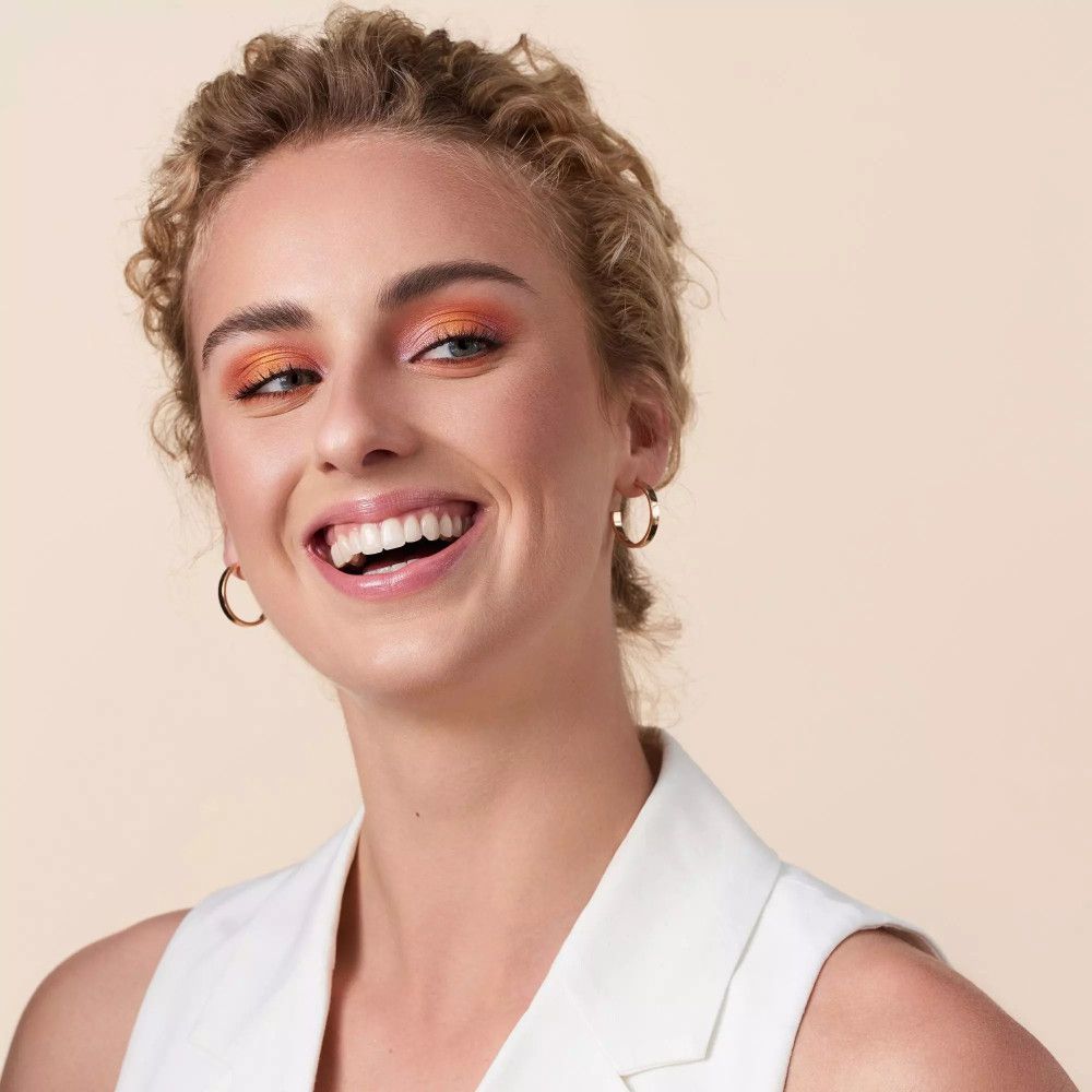 Frau mit lockigem blondem Haar und orangefarbenem Lidschatten, die lächelt. Sie trägt goldene Ohrringe und eine weiße Weste.