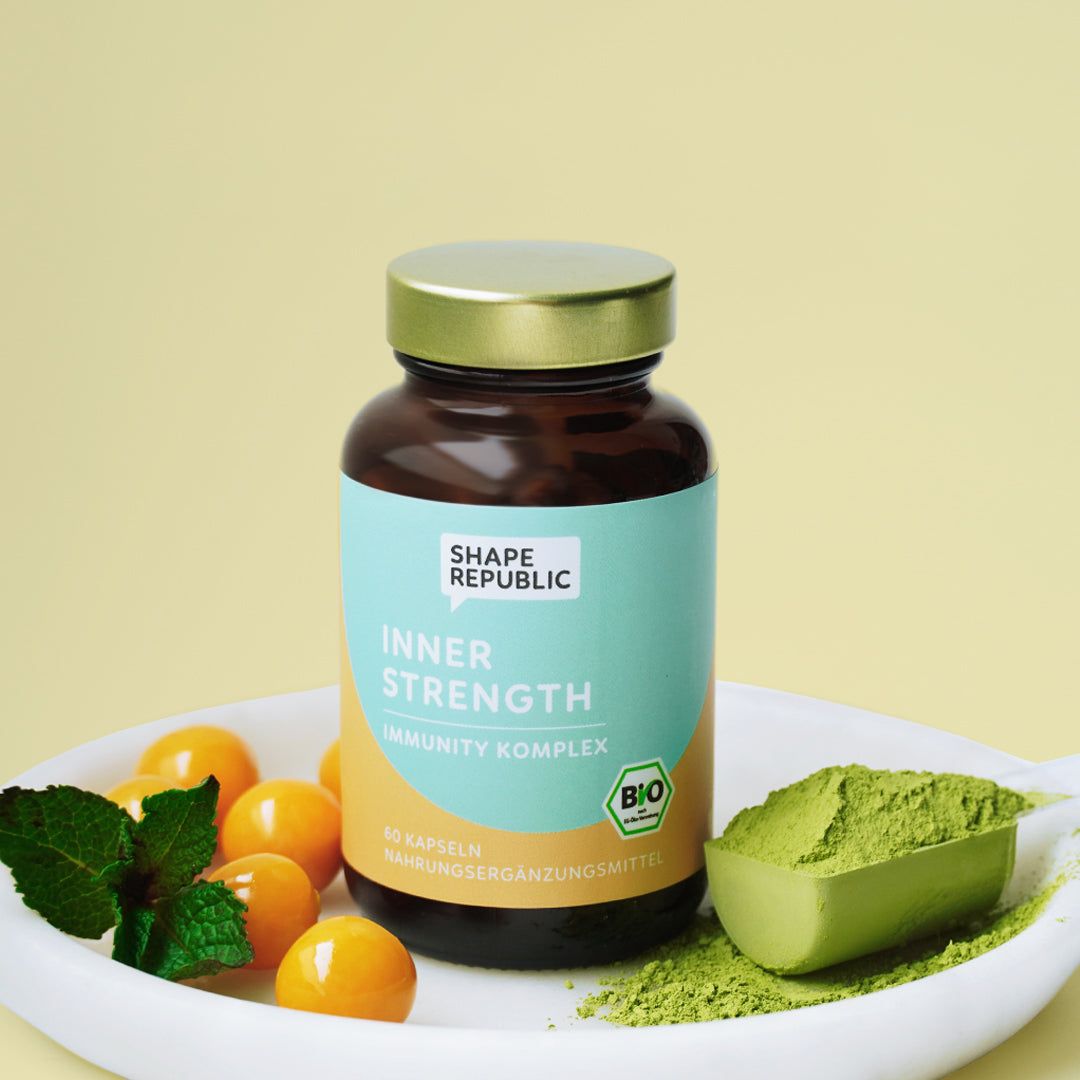 Braune Glasflasche mit Kapseln, daneben Matcha-Pulver und Früchte. Aufschrift: Shape Republic, Inner Strength.