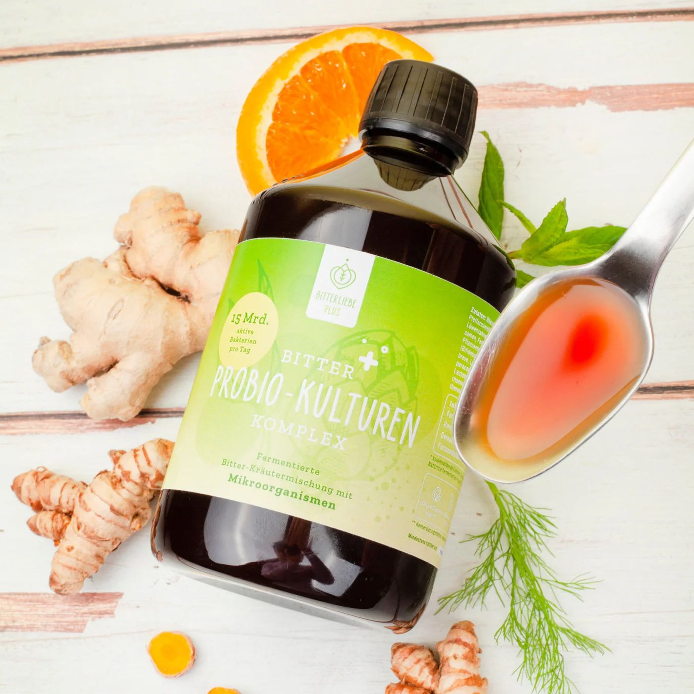 Flasche mit grünem Etikett, daneben Ingwer, Kurkuma, Orange, Löffel mit Flüssigkeit. Aufschrift: Bitter Probio-Kulturen Komplex. 15 Mrd. aktive Kulturen pro Tag.