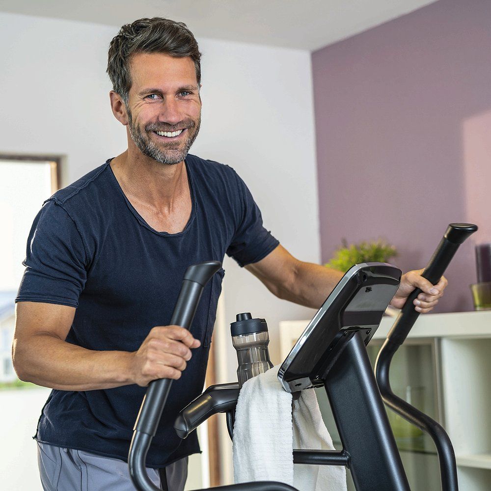 Mann trainiert auf einem Ellipsentrainer. Er lächelt. Im Hintergrund: Zimmer, Flasche, Handtuch.
