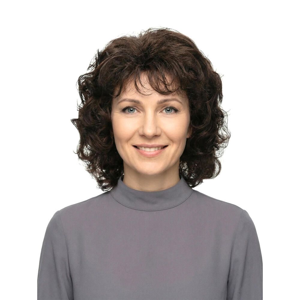 Frau mit brauner, lockiger Perücke. Frontalansicht, lächelnd. Graues Oberteil.