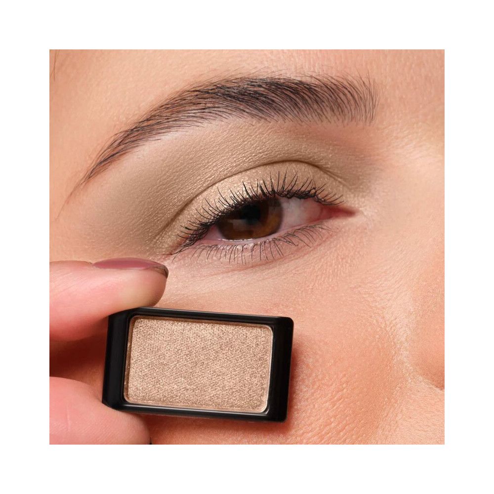 Ein einzelner Lidschatten in einem schwarzen Behälter. Der Lidschatten ist hellbraun und wird vor einem Auge gehalten.