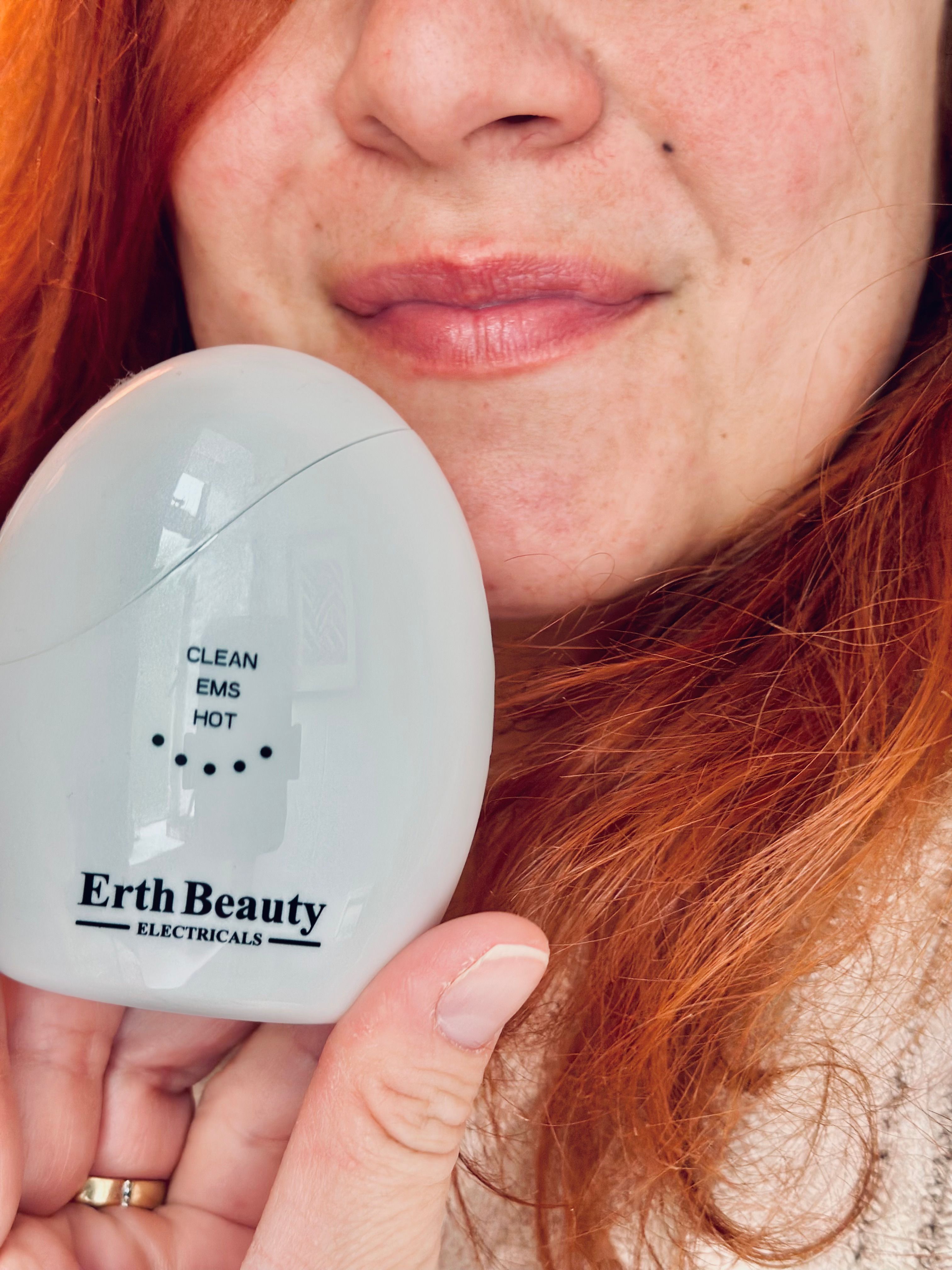 Weißes, ovales Gerät mit Beschriftung: CLEAN, EMS, HOT. Marke: Erth Beauty Electricals. Gerät wird gehalten.