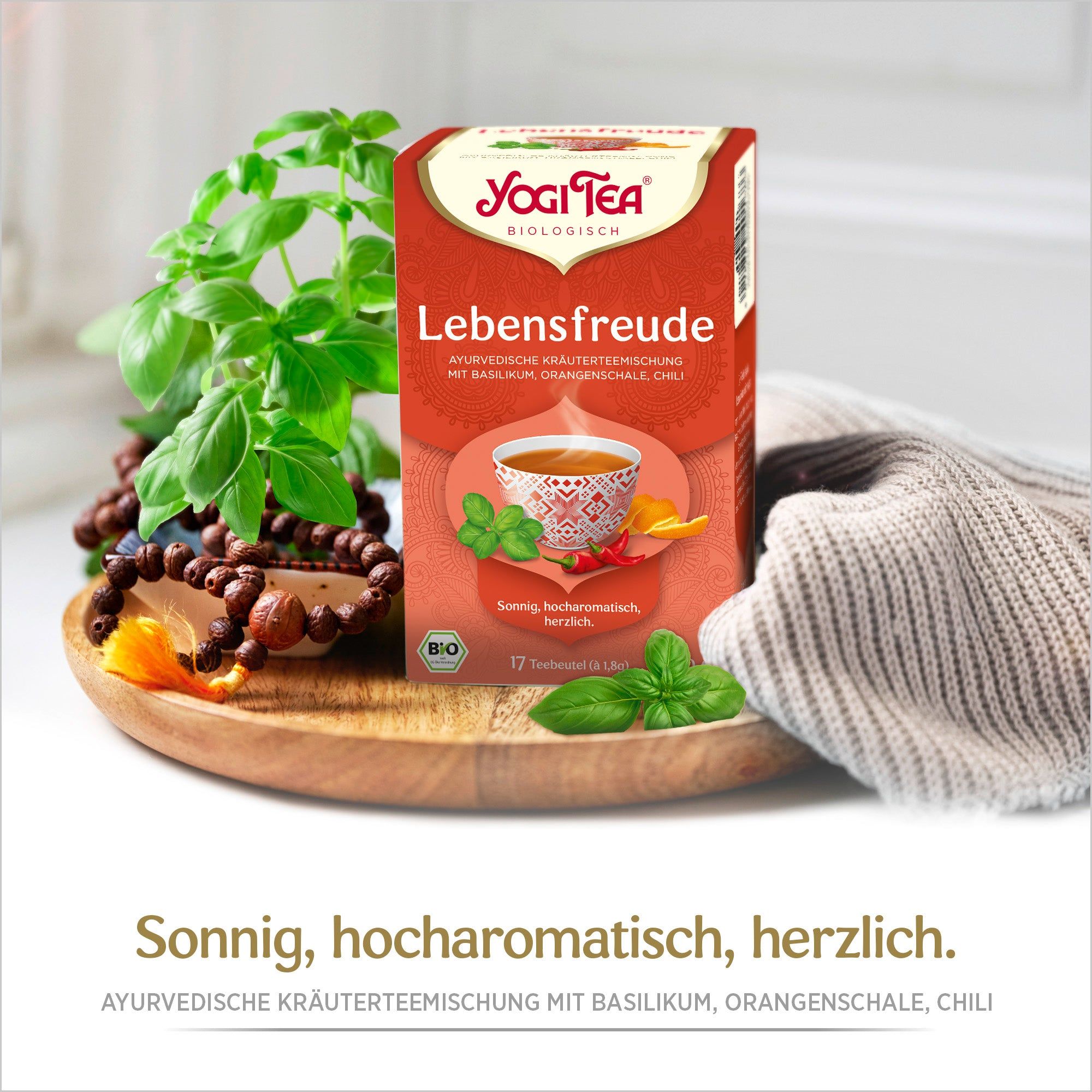 Yogi Tea Lebensfreude Packung. Rote Verpackung mit Logo und Produktinformationen. Bio-Siegel. Basilikum und Holzschale.