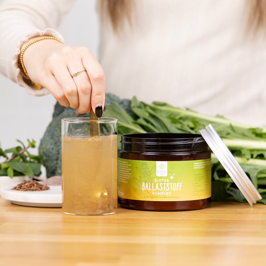 Person rührt Pulver in Glas. Daneben Dose mit gelb-grünem Etikett. Text: BitterLiebe Ballaststoff Komplex. Gemüse und Kräuter im Hintergrund.