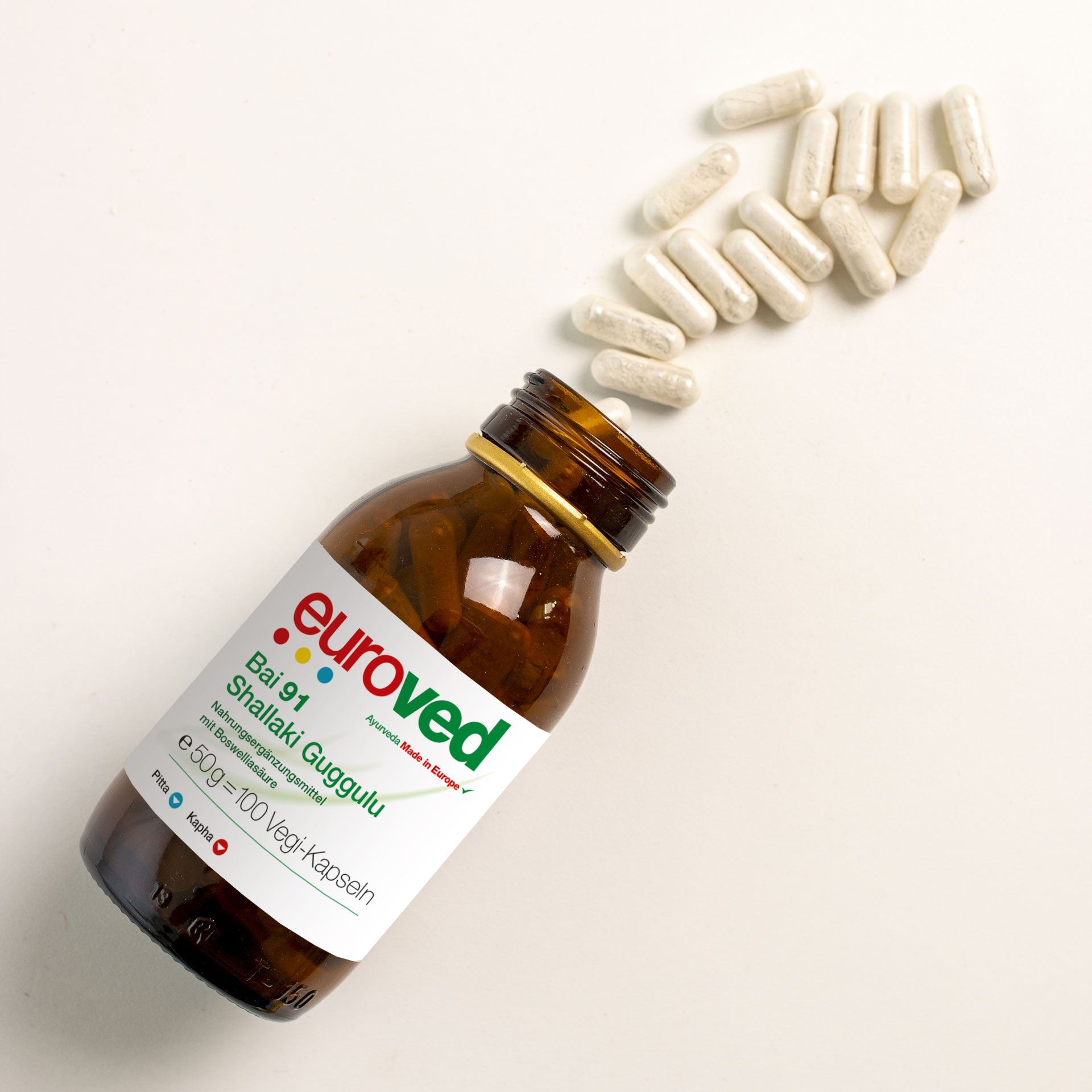 Braune Glasflasche mit weißem Etikett. Kapseln liegen daneben. Aufschrift: euroved Bai 91 Shallaki Guggulu.