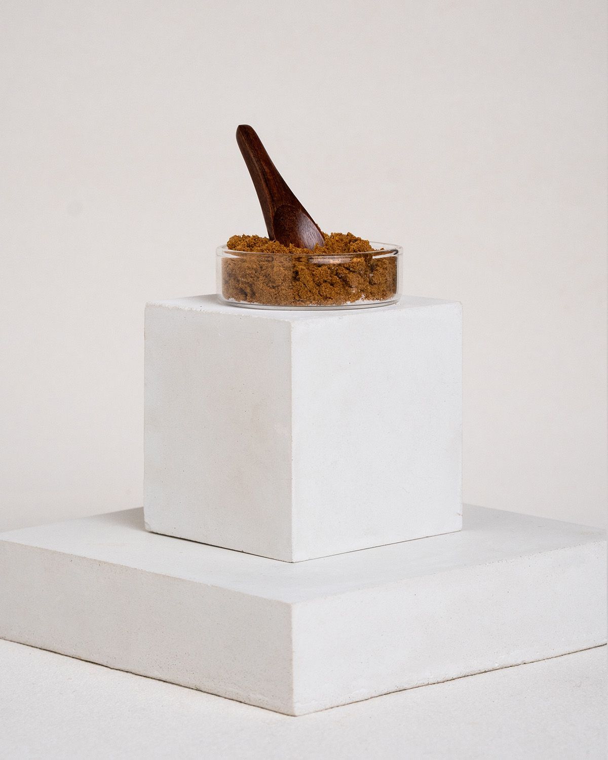 Glasbehälter mit braunem Pulver und Holz-Löffel. Auf weißem Podest platziert.