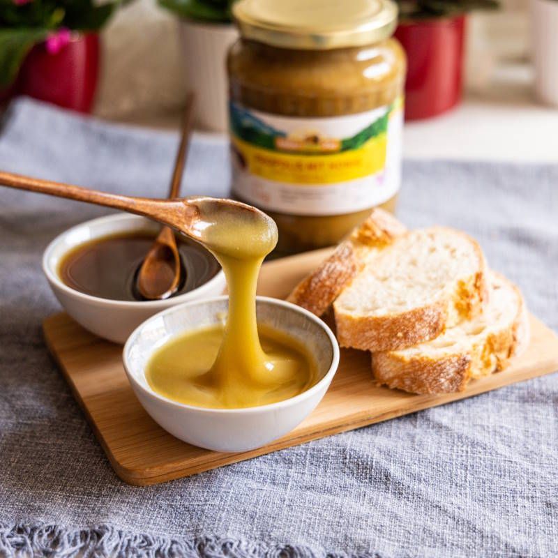 Honig tropft aus einem Holzlöffel in eine Schale. Brot auf Holzbrett. Glas Honig mit Propolis.