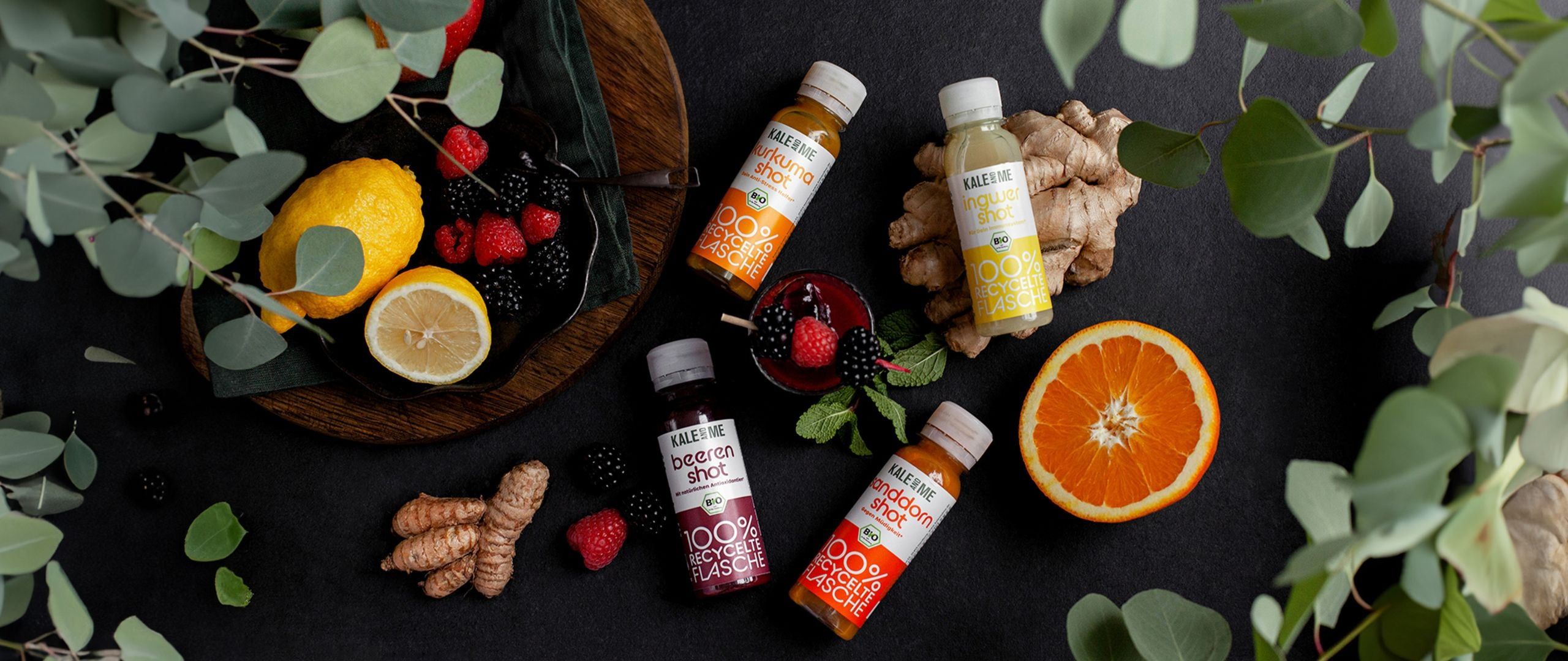 Fünf kleine Flaschen mit Shots, umgeben von Obst und Gemüse. Auf jeder Flasche steht "KALE ME" und der Name des Shots.