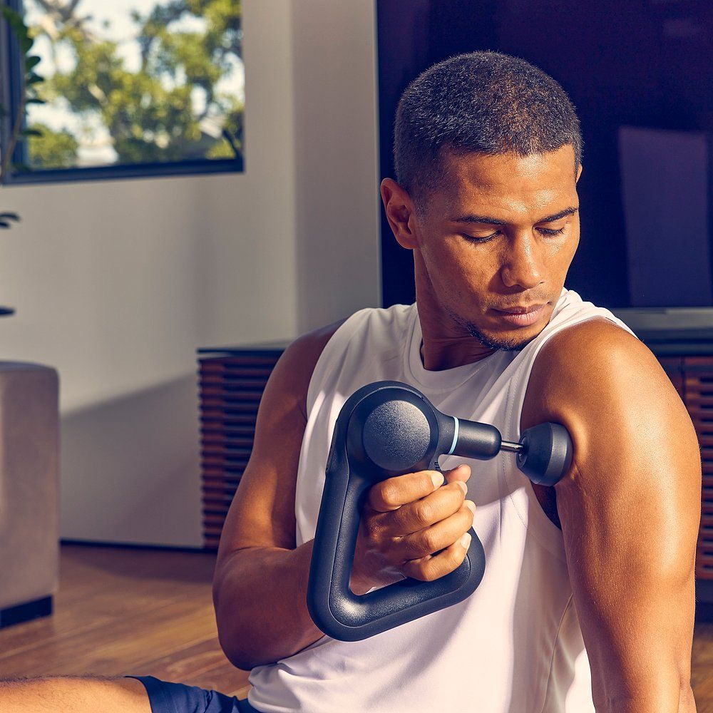 Person verwendet schwarzes Massagegerät am Arm. Dreieckiger Griff. Blaue Akzente. Therabody-Logo.