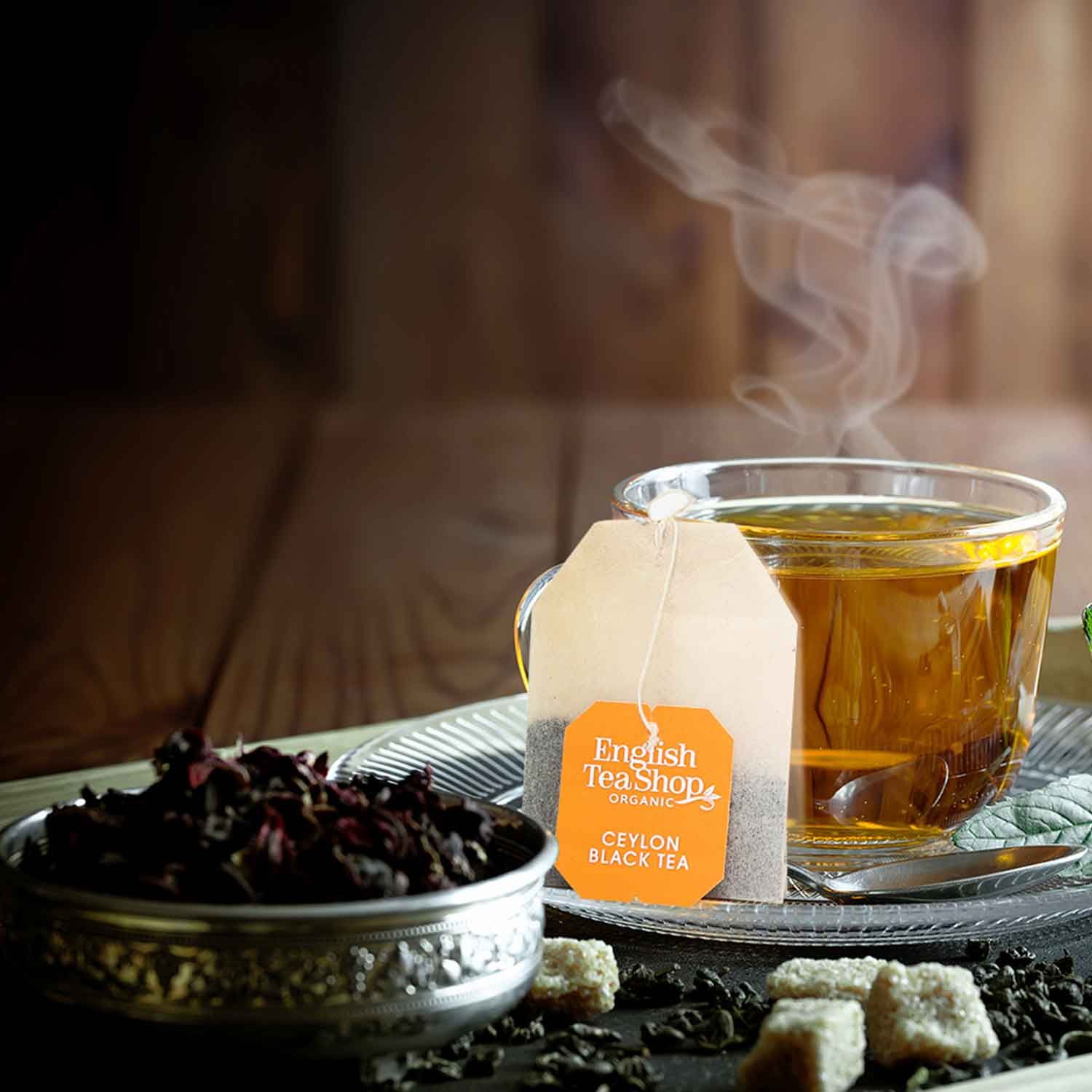 Glas Tee mit Teebeutel "English Tea Shop Ceylon Black Tea". Daneben Schale mit Kräutern und Zuckerwürfel.