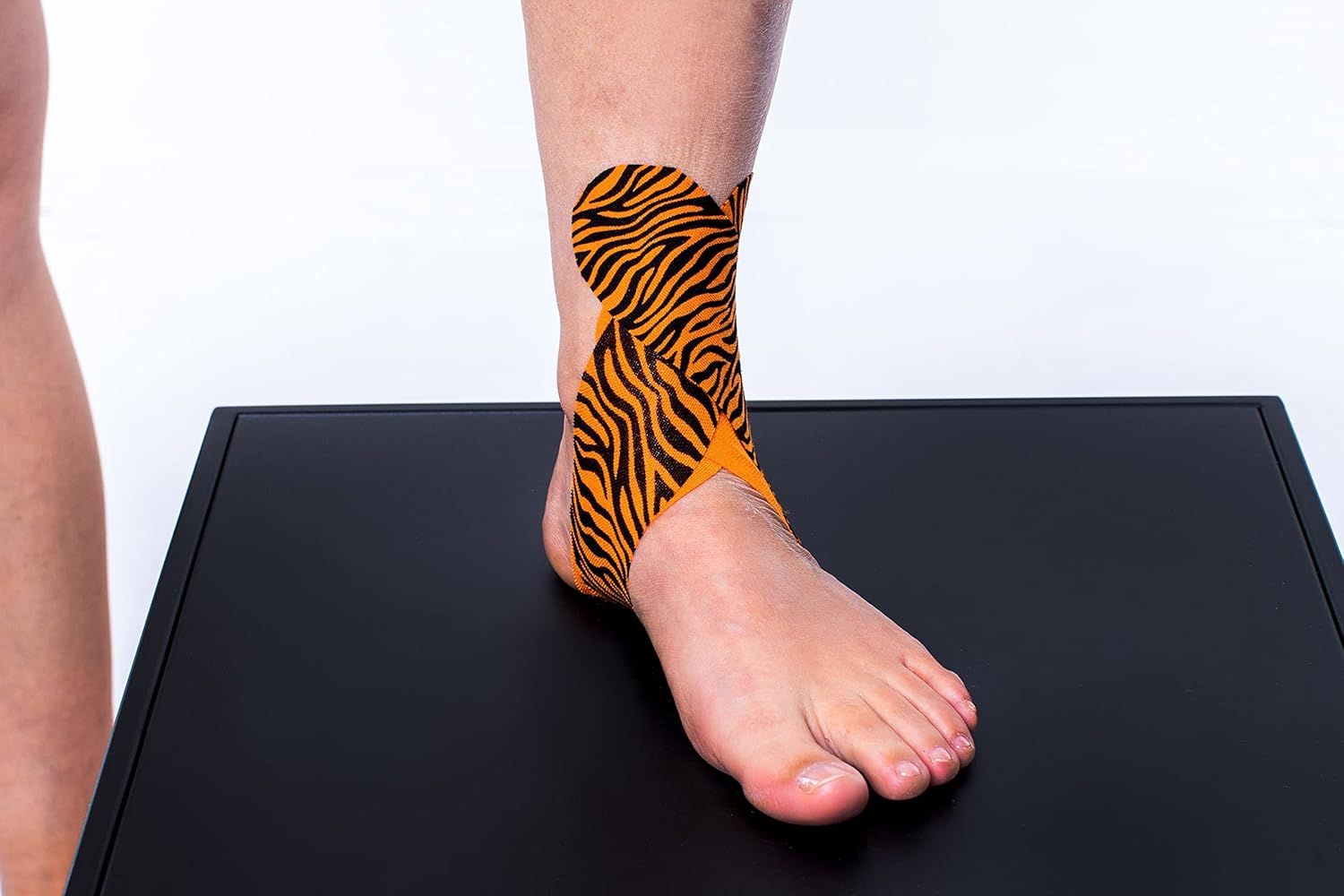 Fuß und Knöchel mit CureTape Tiger. Orange mit schwarzen Streifen. Auf schwarzem Podest. Anwendung am Knöchel.