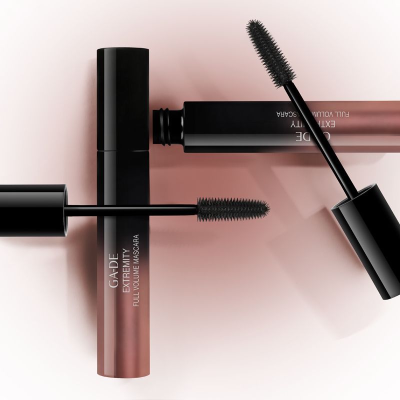 Drei Mascara-Tuben. Zwei mit herausgezogenen Bürsten. Aufschrift: GA-DE Extremity Full Volume Mascara. Rosa Hintergrund.