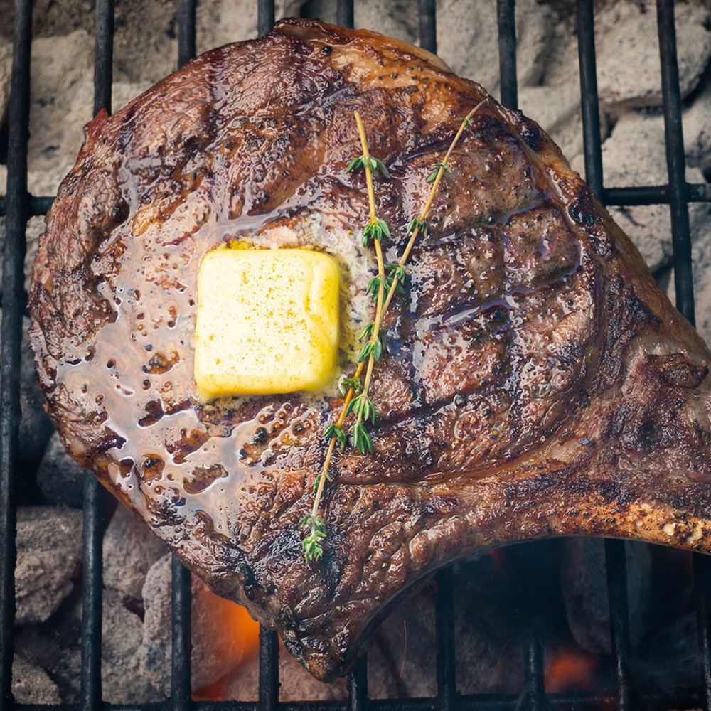 Gegrilltes Steak mit Butter und Kräutern. Auf einem Grillrost. Fleisch braun, Butter gelb, Kräuter grün. Holzkohle im Hintergrund.