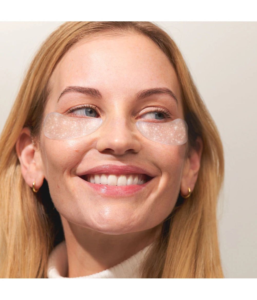 Vrouw met oogpads op haar ogen. Pads zijn transparant met glitter. Ze lacht.