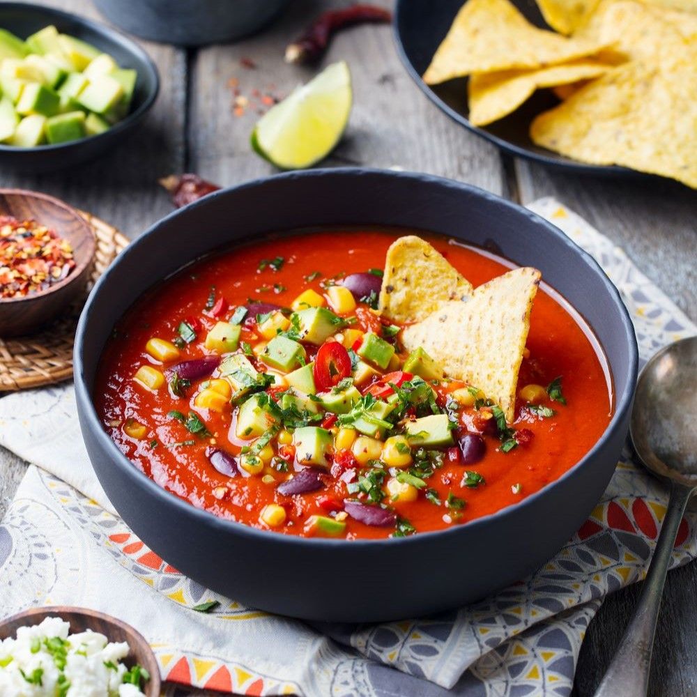 Schwarze Schüssel mit roter Suppe, Avocado, Mais, Bohnen, Tortilla-Chips. Auf Holztisch, weitere Zutaten.