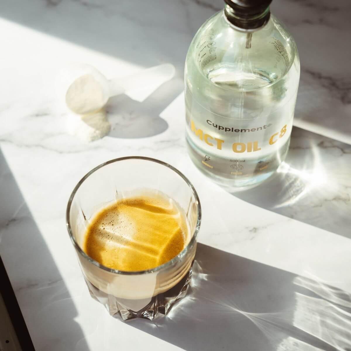 Glas mit Getränk, daneben Flasche mit Etikett "Cupplement MCT OIL C8". Löffel auf Marmoroberfläche.