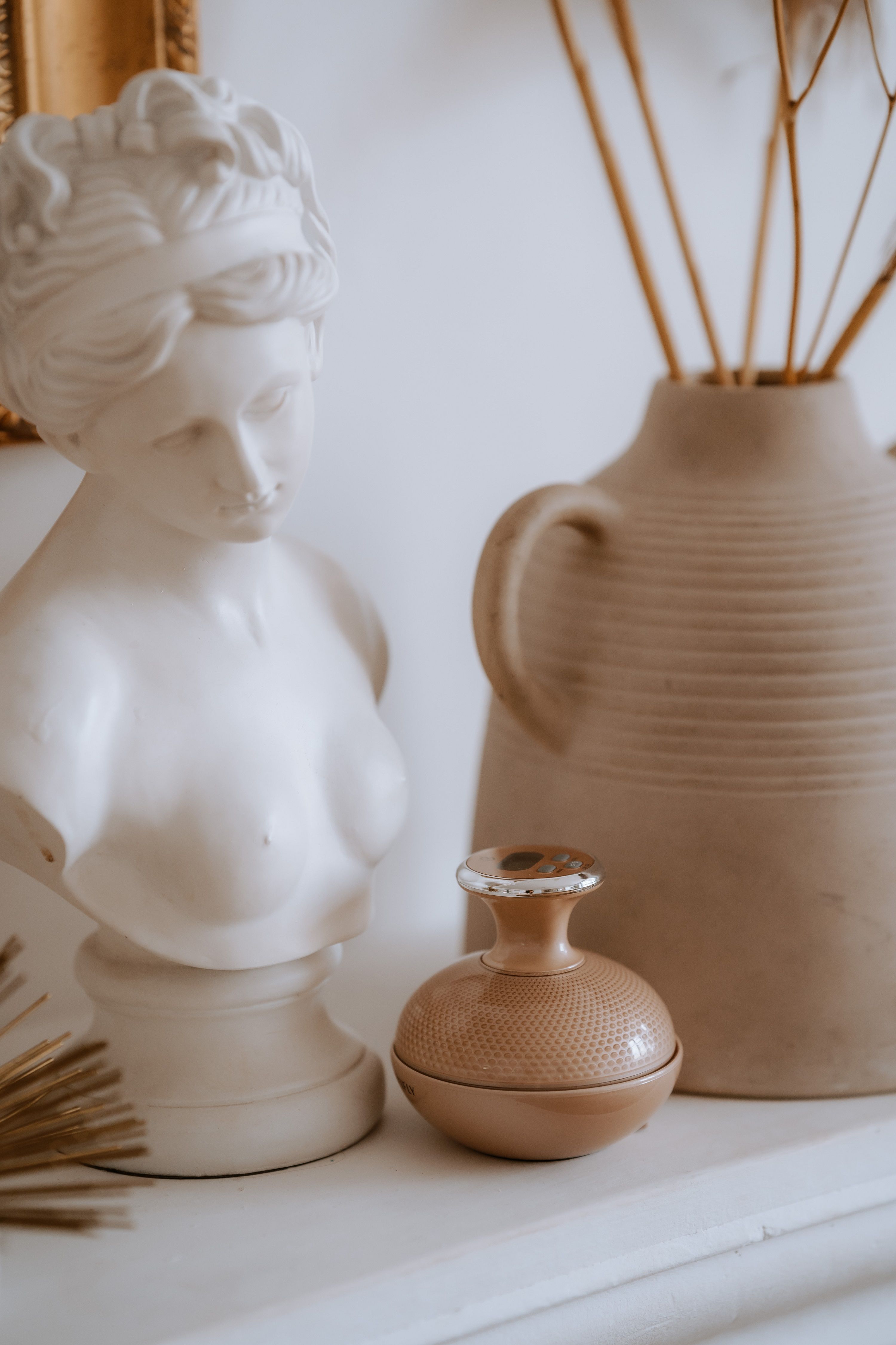 Beige Massagegerät neben Büste und Vase. Gerät mit Perlenstruktur und Griff.