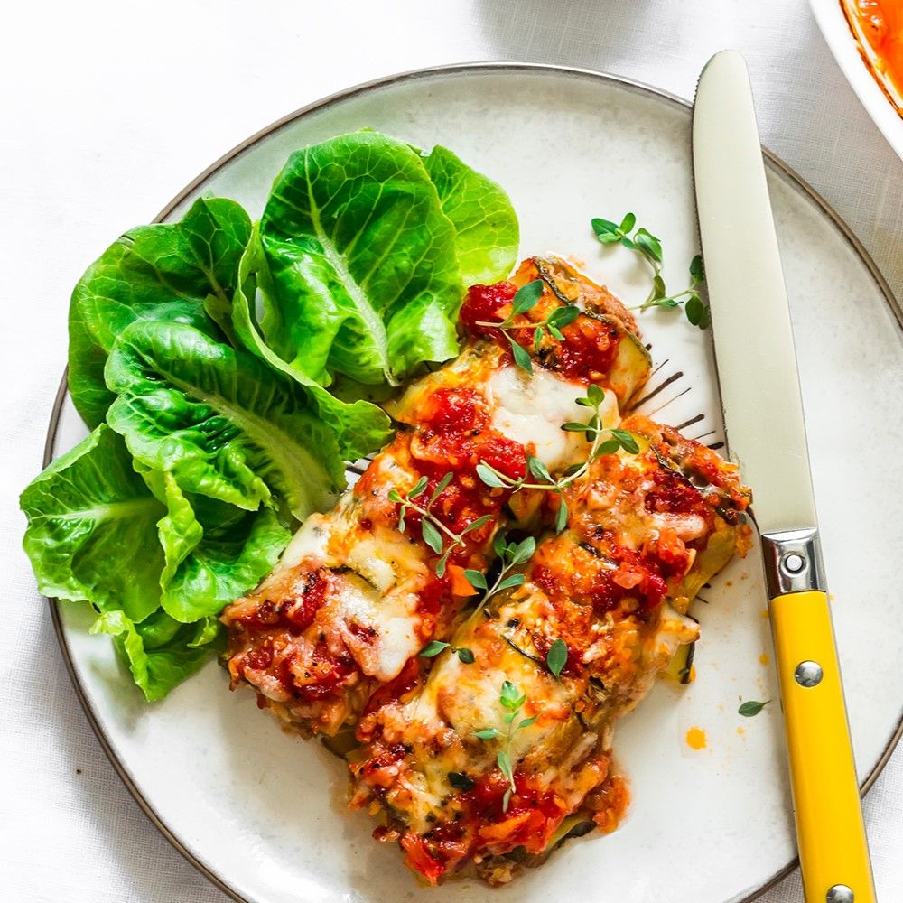 Teller mit Zucchiniröllchen, Tomatensauce, Salat und Messer.