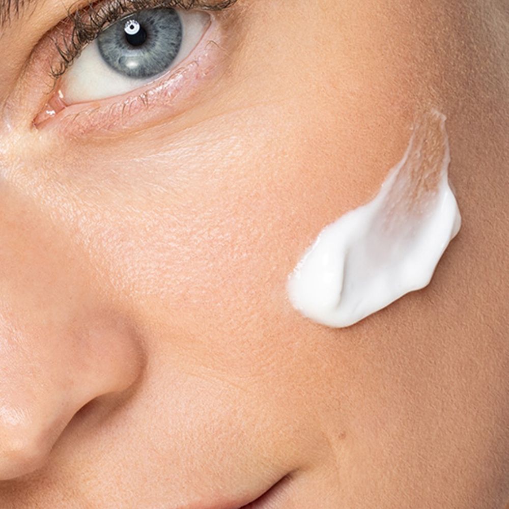 Nahaufnahme eines Gesichts mit einem Klecks weißer Creme auf der Wange. Blaue Augen, helle Haut.