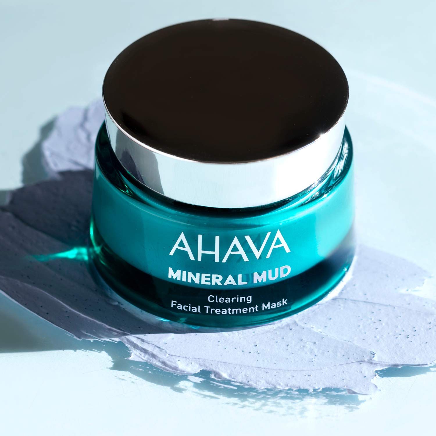 Ahava Mineral Mud Maske. Türkisgrünes Glas, silberner Deckel. Produktname und Text sichtbar. Auf Untergrund.