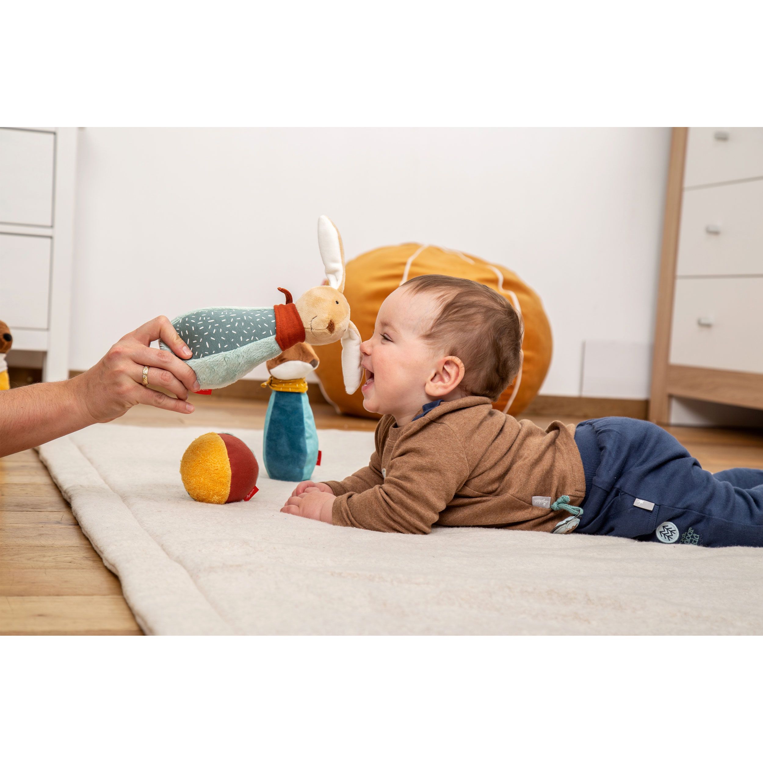 Baby spielt mit Kegelspiel-Figuren. Hase und Kugel sind sichtbar. Auf einer Decke. Person hält Figur.