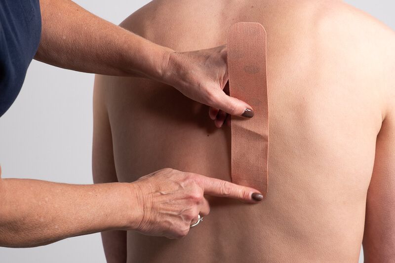 Person zeigt Kinesiotape-Streifen auf dem Rücken einer Person. Tape ist beige.