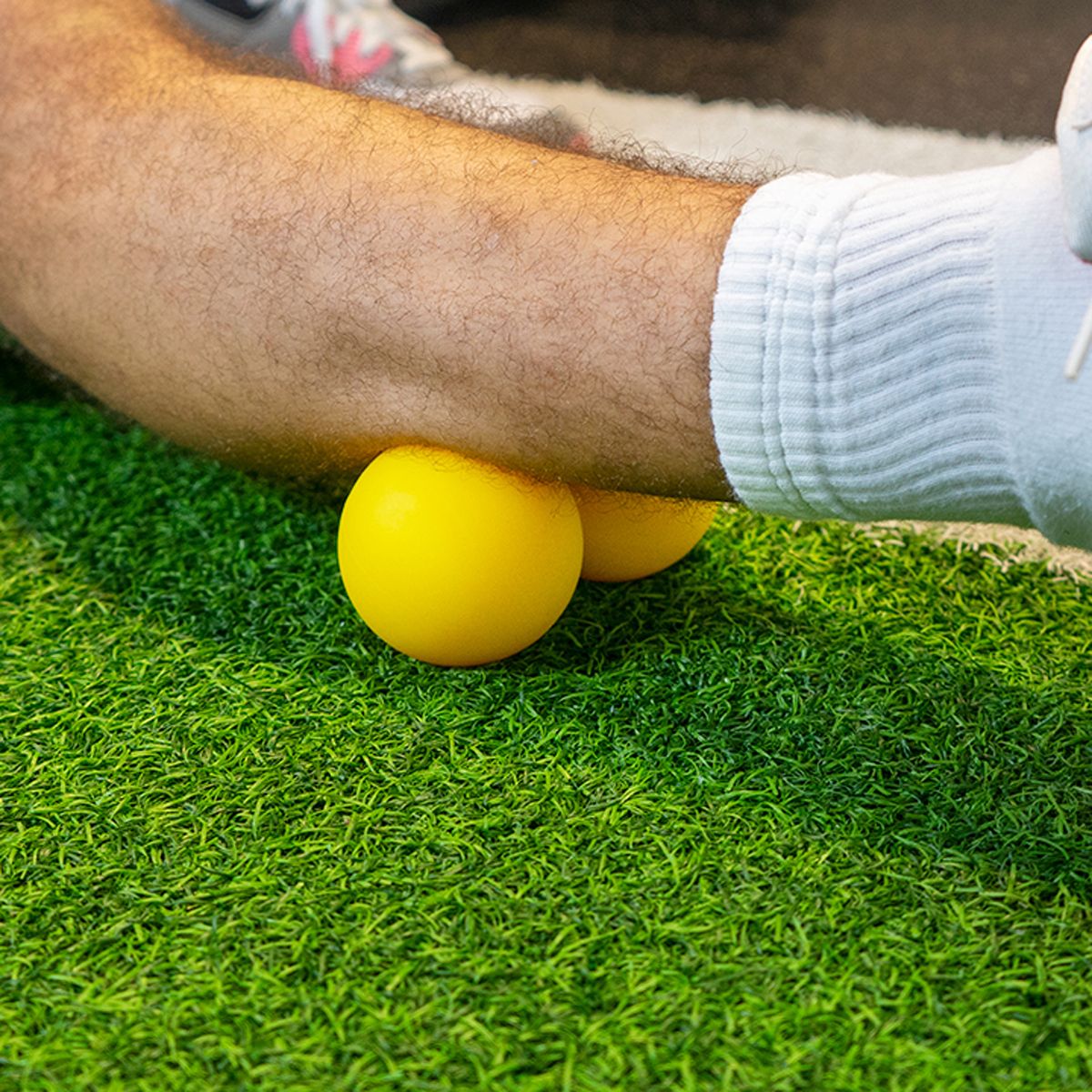 Gelber Doppel-Massageball unter Arm. Auf grünem Kunstrasen. Person trägt weiße Socken und Sportschuhe.