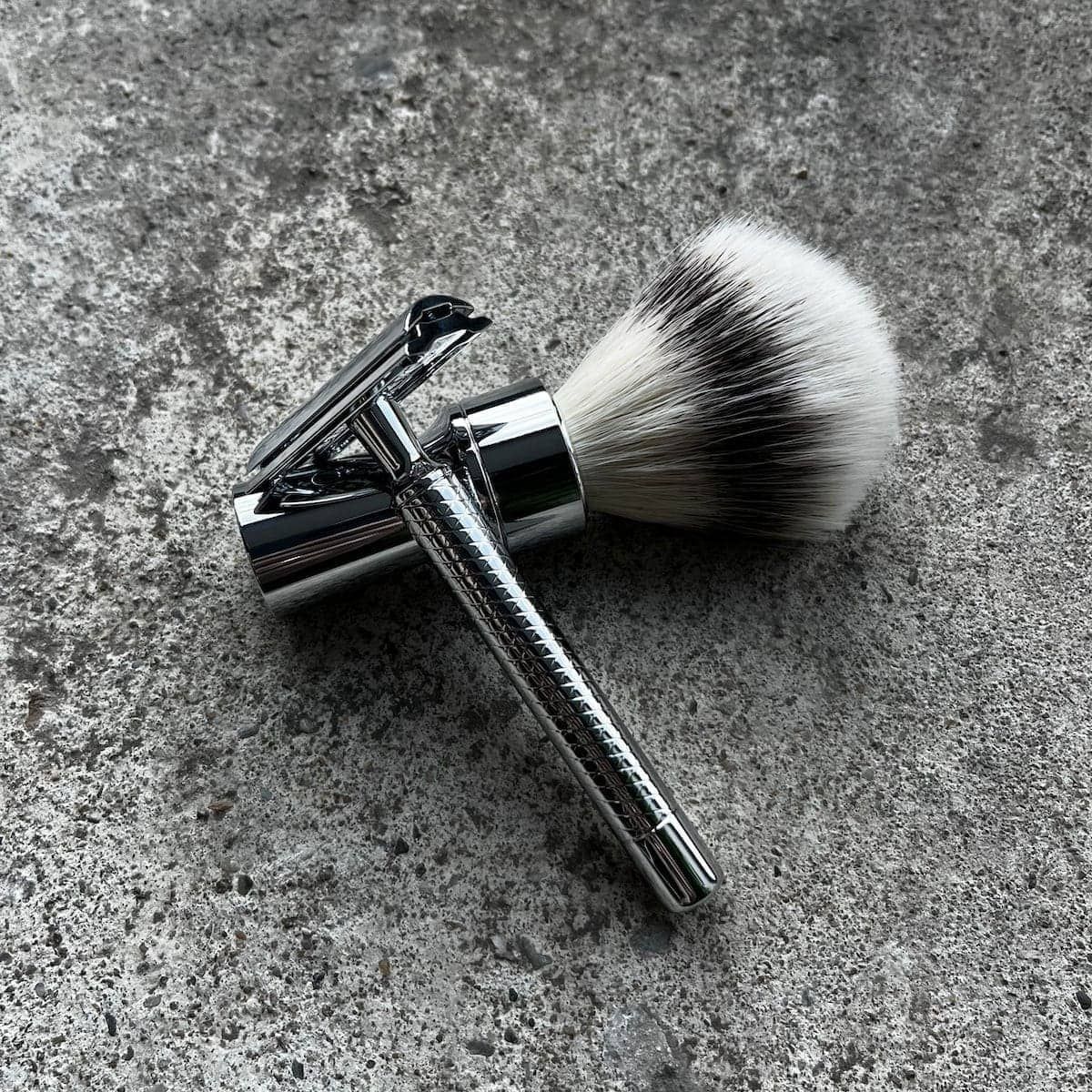 Rasierpinsel mit weiß-schwarzen Borsten und Rasierer mit geriffeltem Griff. Beide aus Metall, auf grauem Untergrund.