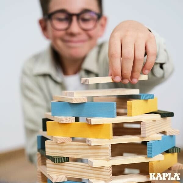 Kind baut Turm aus Holzplanken. Einige Planken sind blau und gelb gefärbt. Aufschrift: KAPLA.