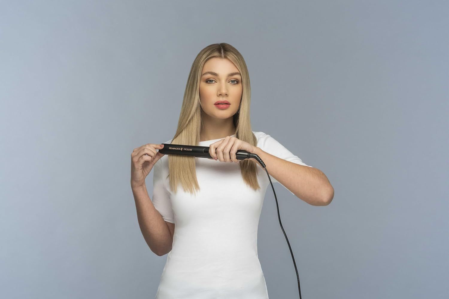 Blonde Frau hält ein schwarzes Glätteisen. Sie blickt in die Kamera. Das Glätteisen ist in Gebrauch.