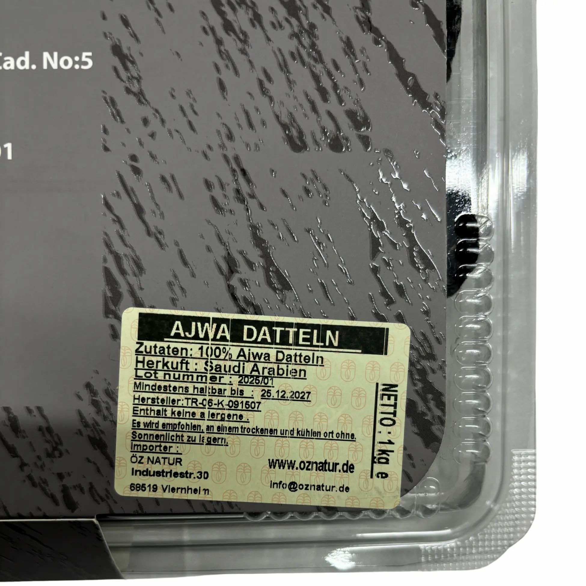 Nahaufnahme der Verpackung mit Etikett. Aufschrift: AJWA DATTELN, Zutaten, Herkunft, Mindesthaltbarkeit, Hersteller, Importeur.