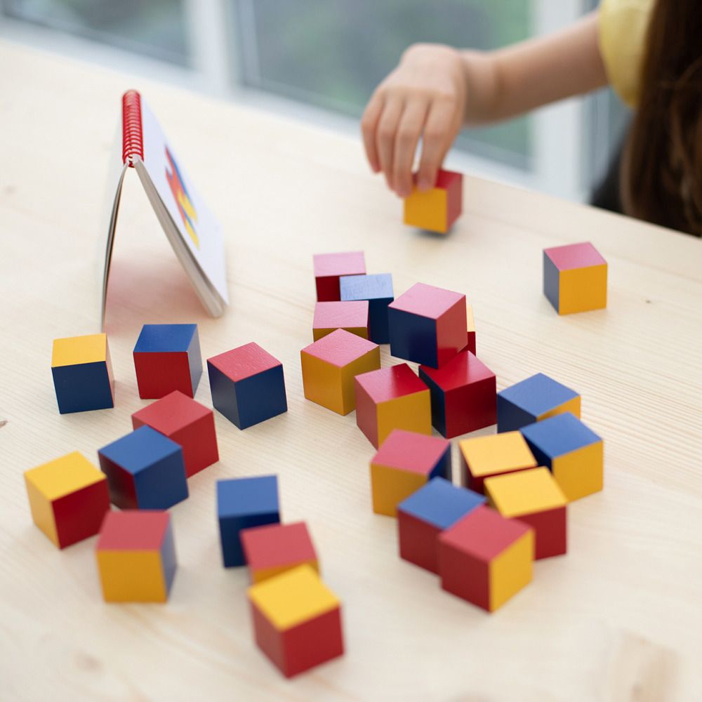 Bunte Würfel auf einem Holztisch. Gelbe, rote und blaue Würfel. Ein Heft mit Vorlagen liegt daneben. Kind greift nach einem Würfel.