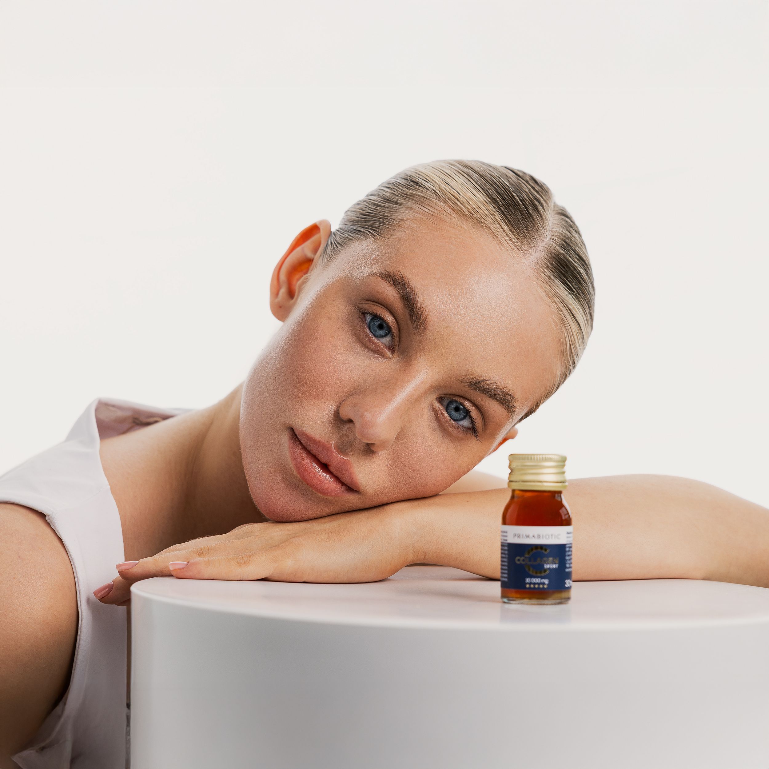 Frau lehnt an einer weißen Säule. Hält eine Flasche Primabiotic Collagen Sport. Goldfarbener Deckel.